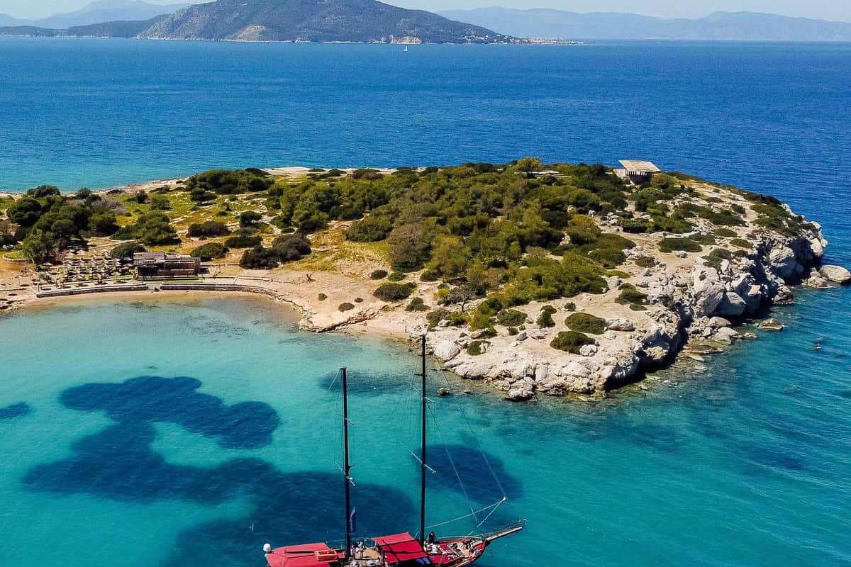 Aegina boat trip wooden two-masted boat anchored in turquoise water beside the pine-covered islet of Moni