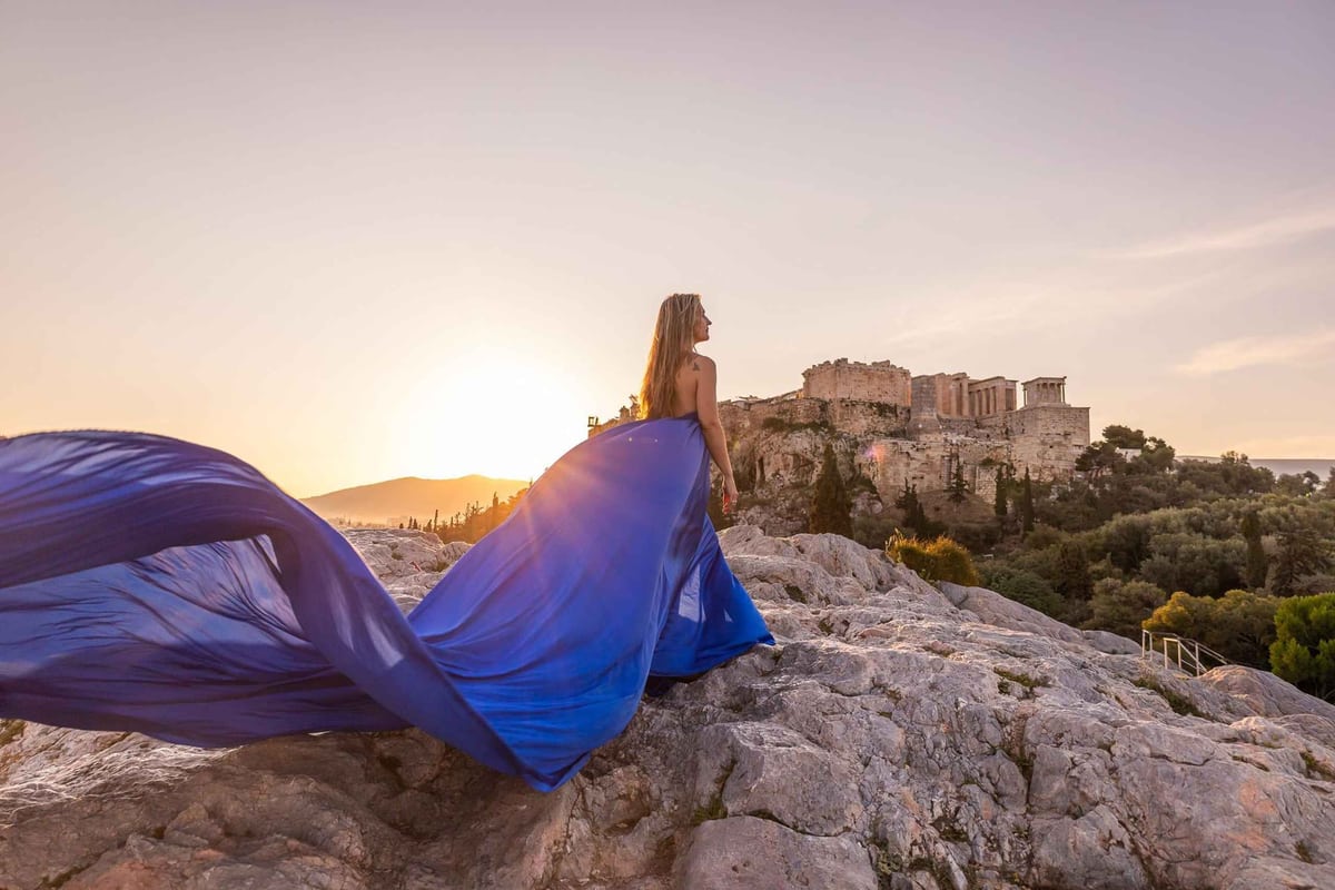 An Athens photo tour poses a traveler in a flowing dress on the rocks facing the Acropolis in the evening light