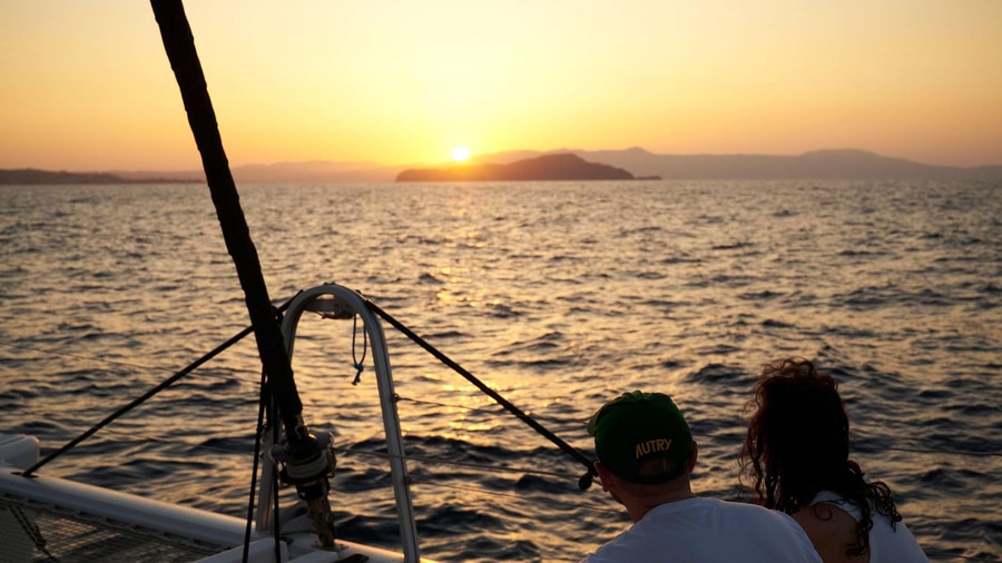 Two passengers on the net of a sailing catamaran watch the sun set behind an islet during a Chania catamaran cruise
