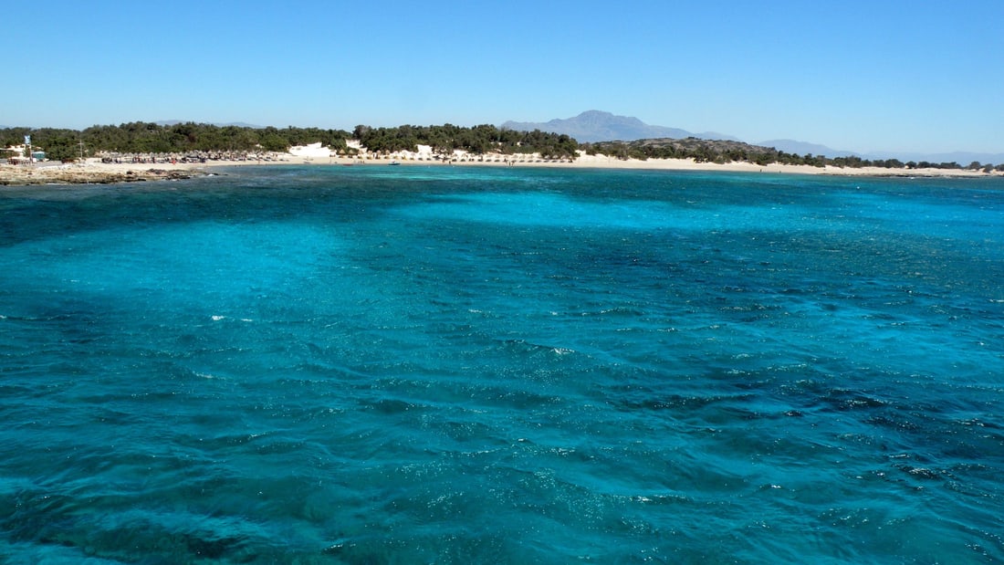 A Chrissi island cruise crosses clear turquoise water toward the white beach and juniper trees of Chrissi