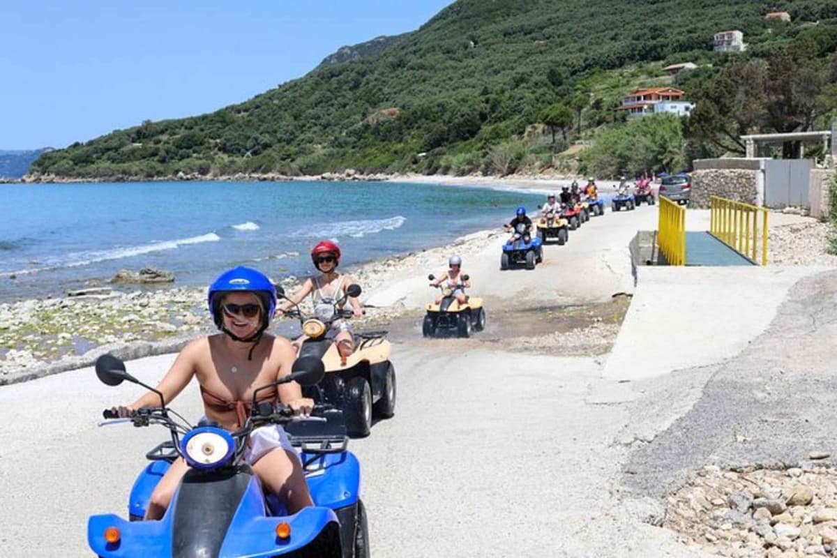 A Corfu ATV tour line of quad bikes rides a coast road beside a blue bay and green hills