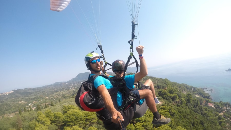 A pilot and passenger in helmets fly a tandem wing above green wooded hills during Corfu paragliding