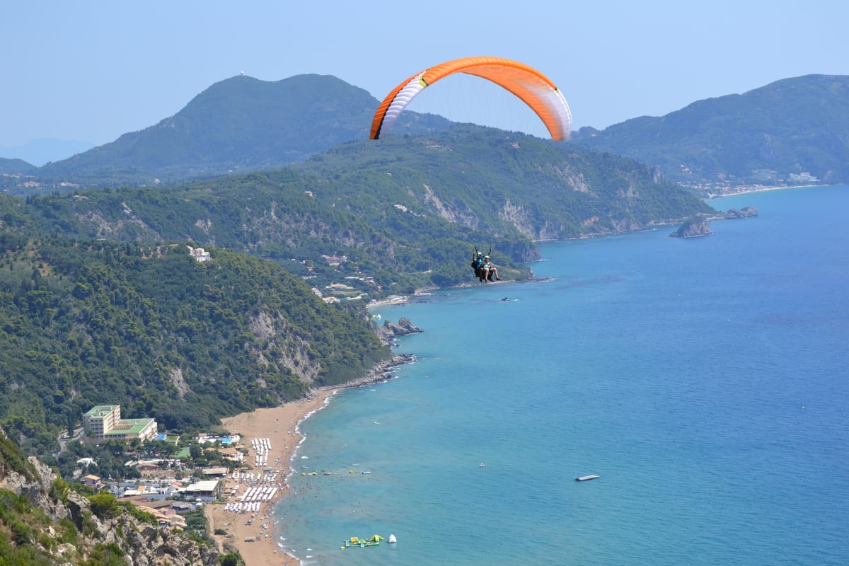 Corfu paragliding orange wing high above the green west coast of Corfu and a long sandy bay