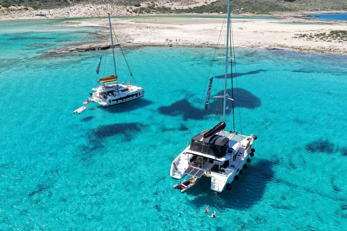 A Crete catamaran cruise anchors two sailing catamarans in the shallow turquoise water of Balos lagoon below a sandy spit