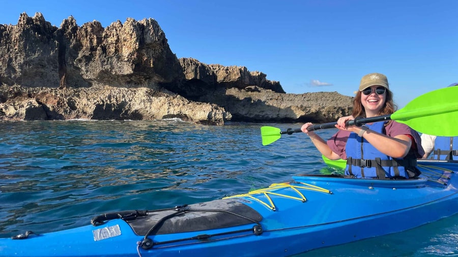 A Crete kayak tour paddler smiles in a blue sea kayak below jagged rocks on the Chania coast