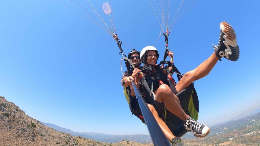 Crete paragliding tandem pair in helmets under an orange wing high above dry hills