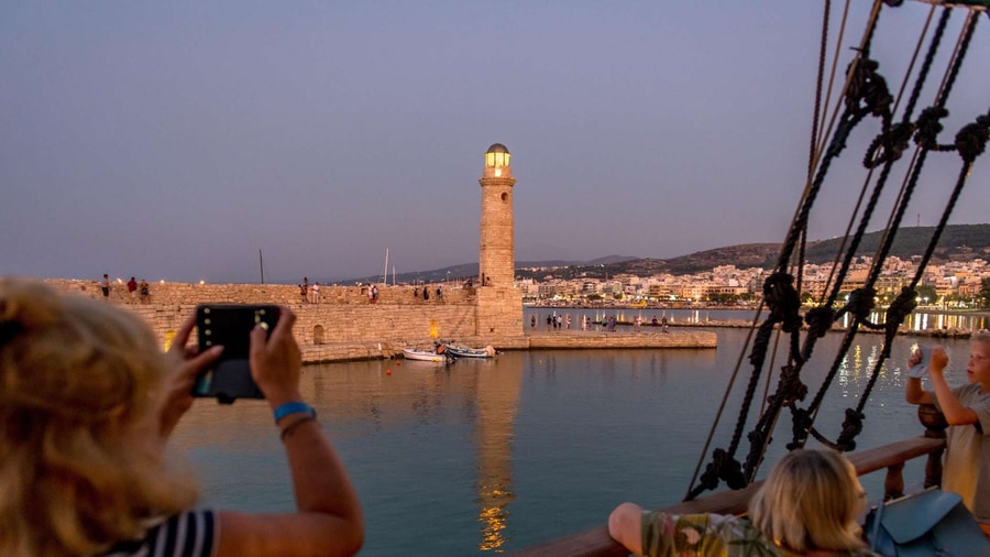 Passengers on a wooden boat photograph the Rethymno harbour lighthouse at dusk during a Crete sunset cruise