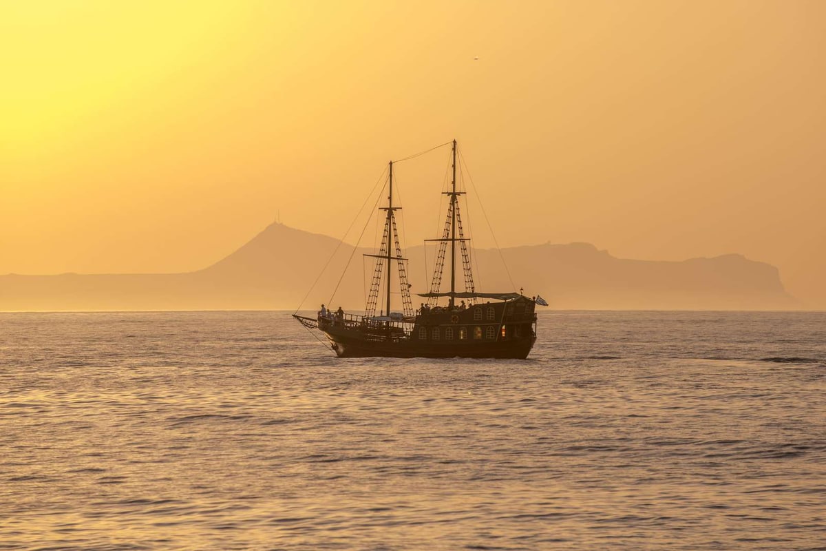 Crete sunset cruise wooden two-masted boat sails past a headland as the sun sets over a golden sea