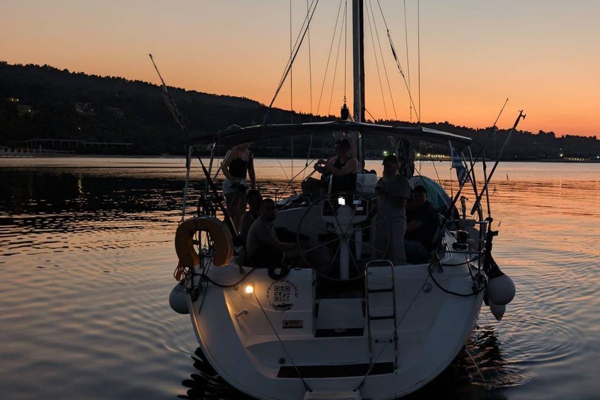 A sailing yacht with guests on deck under an orange sky at sunset off Kassandra on a Halkidiki sunset cruise