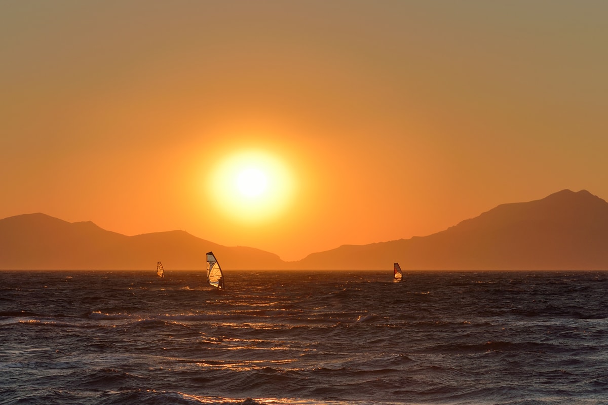 Windsurfers sail under an orange sun setting behind island mountains seen from Kos on a Kos sunset cruise