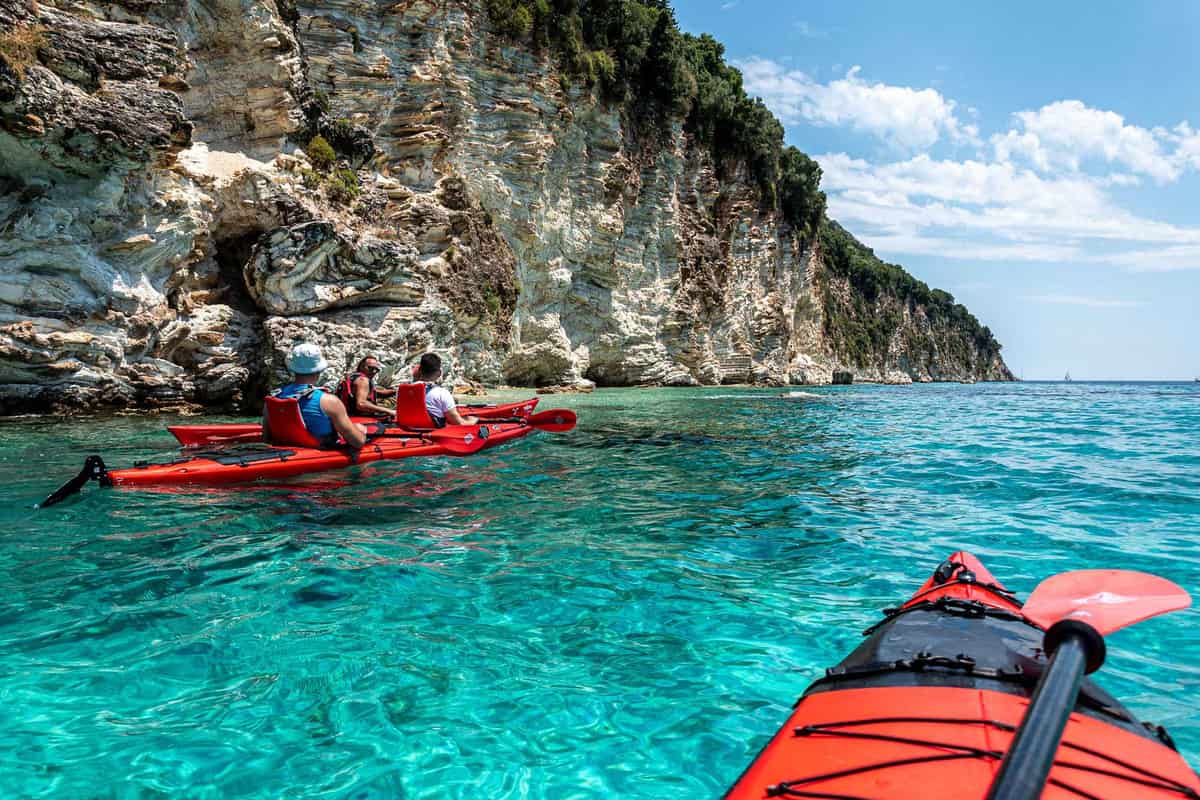 A Lefkada kayak tour paddles red sea kayaks through turquoise water below white limestone cliffs