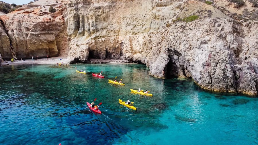 A Milos kayak tour paddles red and yellow sea kayaks across a turquoise cove below pale volcanic cliffs