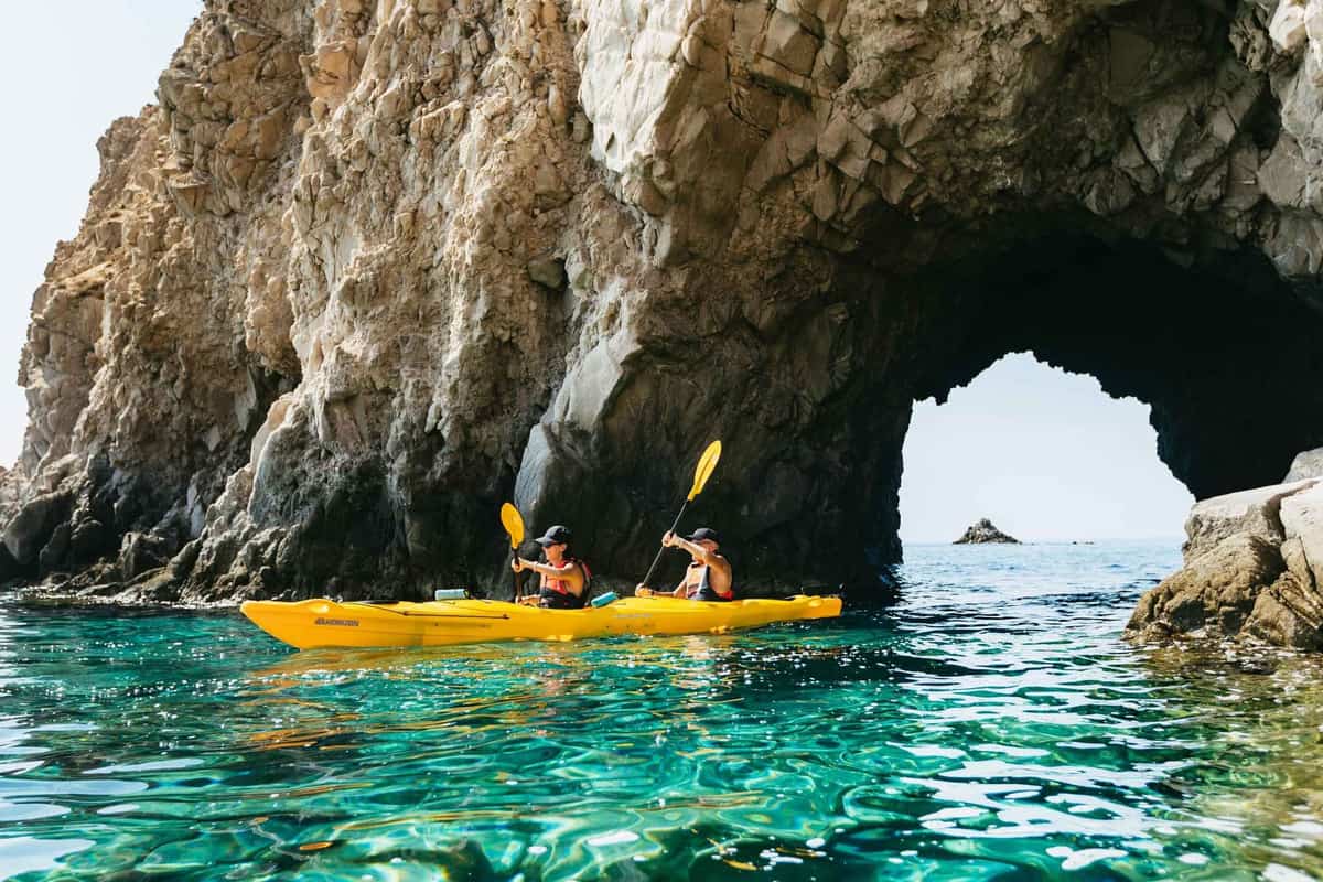 A Milos kayak tour paddles a yellow double kayak beneath a rock arch over clear turquoise water