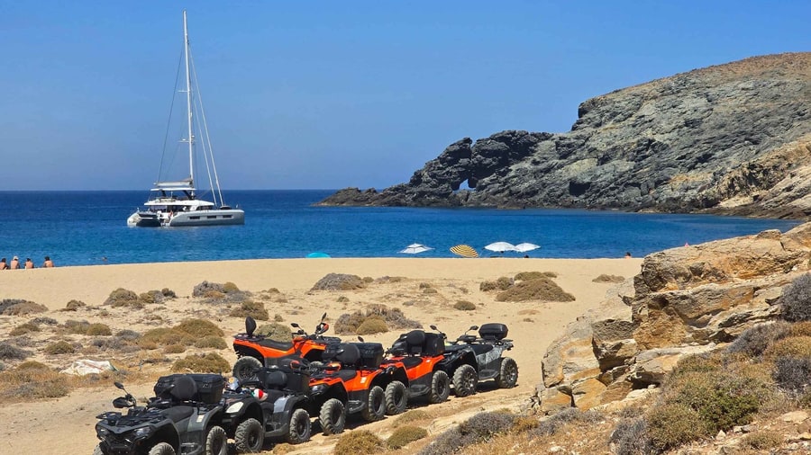 A Mykonos ATV tour parks a line of quad bikes on a sandy northern beach with a sailing boat anchored in the bay