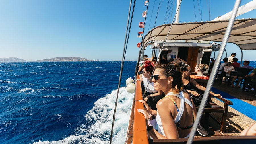 Passengers at the rail of a wooden boat on a Mykonos boat trip, with white wake and blue sea behind
