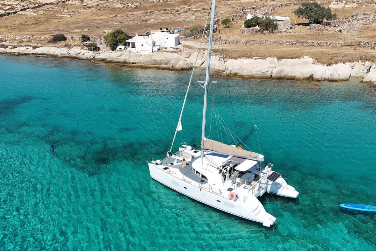 Mykonos catamaran cruise boat anchored in turquoise water below a rocky shore with whitewashed houses