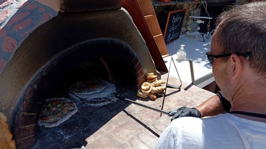 A Mykonos cooking class bakes flatbreads in a wood-fired oven on a farm, with a peel lifting them from the coals