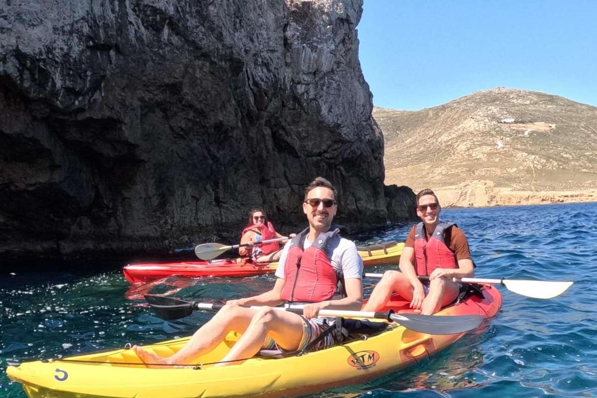 Paddlers in sit-on-top sea kayaks below a steep rock cliff on a Mykonos kayak tour