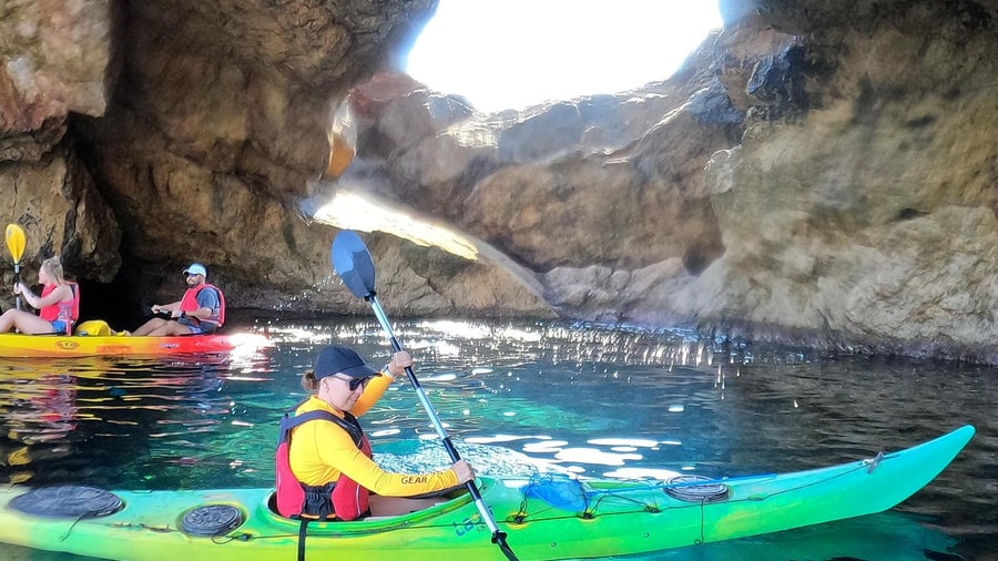 Sea kayakers paddle inside a sea cave lit through an opening in its roof on a Mykonos kayak tour