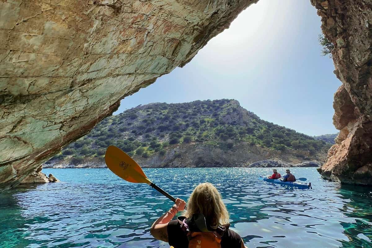 Naxos kayak tour paddler with a yellow paddle under a sea cave arch, with two kayakers and a green hill ahead
