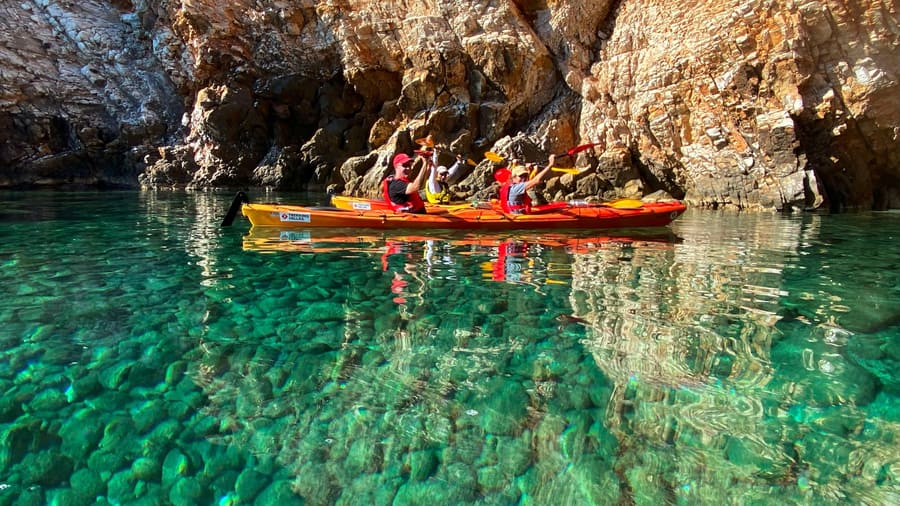 Paddlers in a red tandem kayak float over clear green water below orange cliffs on a Naxos kayak tour