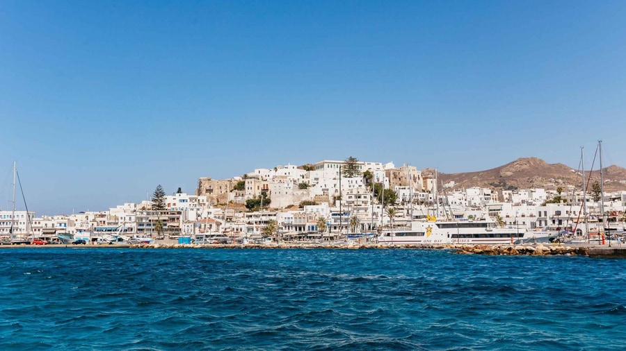 A Naxos sailing trip looks back from the water at the white houses and castle hill of Naxos Town above the harbour
