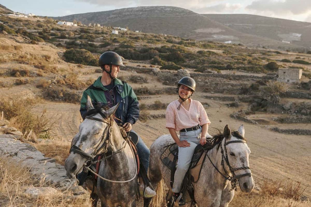 Paros horse riding in the hills, with two smiling riders in helmets on grey horses above dry stone terraces