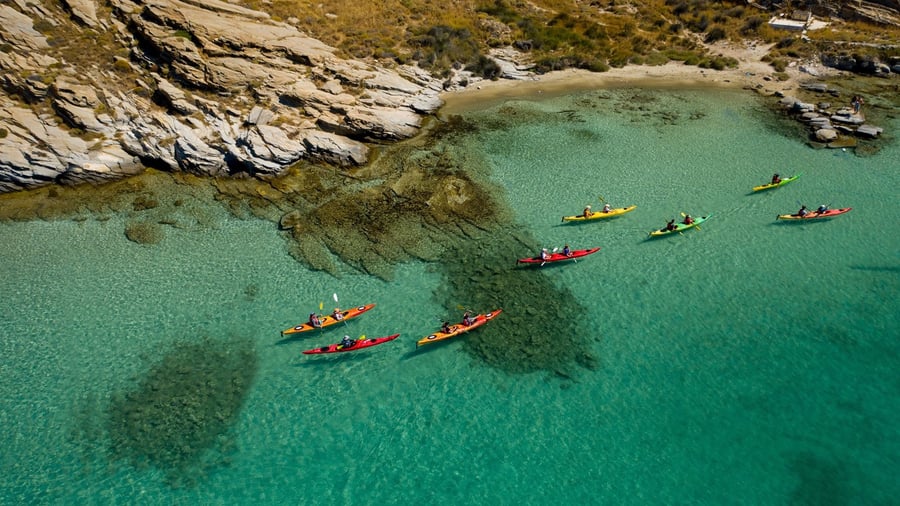 Red and yellow sea kayaks cross a turquoise cove with dark seagrass patches on a Paros kayak tour
