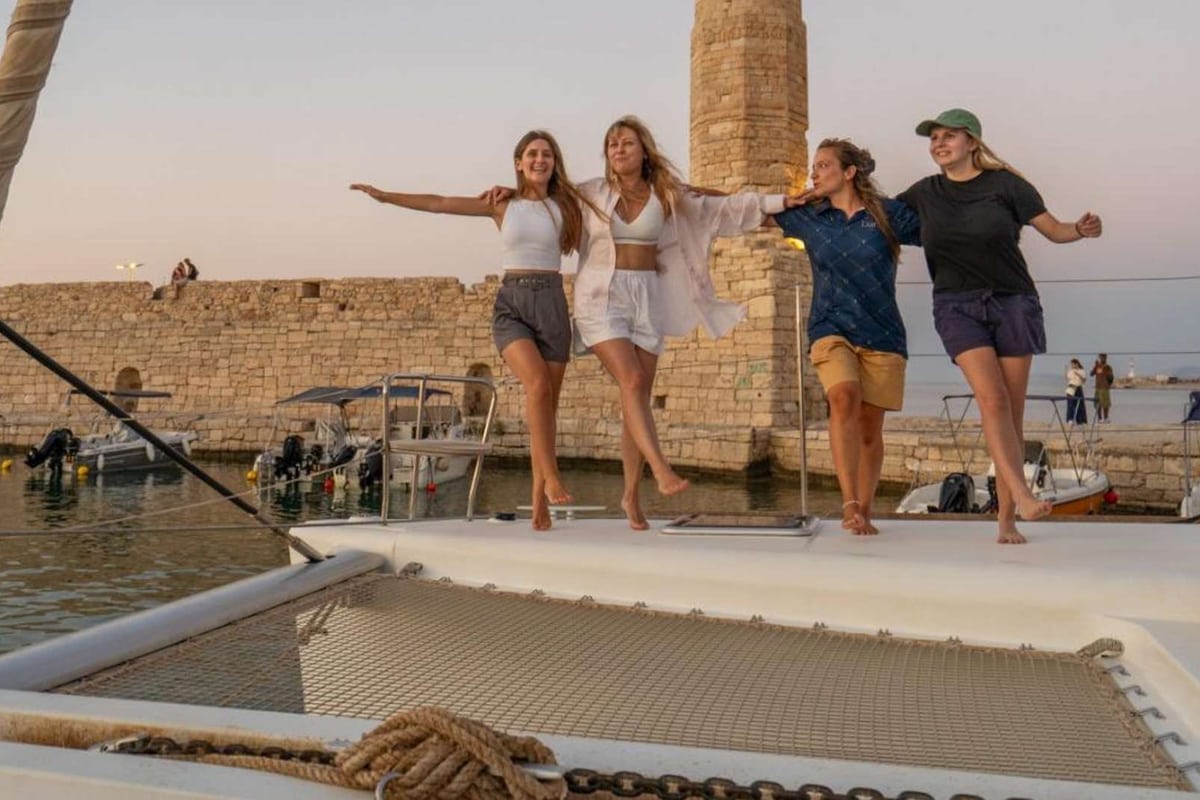 Guests on the deck of a sailing catamaran beside the Egyptian lighthouse of the Venetian harbour on a Rethymno sunset cruise