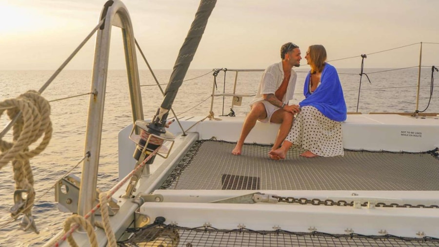 Two guests sit on the net of a sailing catamaran as the sun sets over the sea on a Rethymno sunset cruise