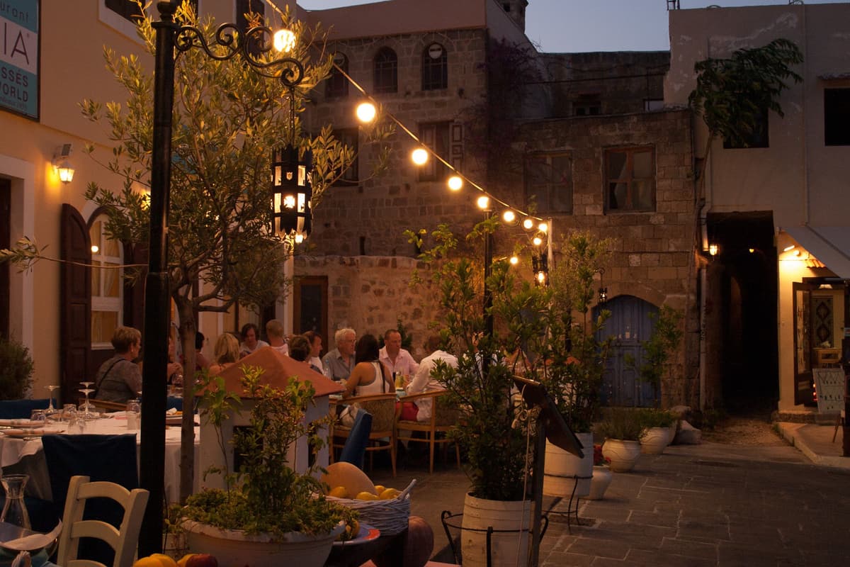 A Rhodes food tour passes tables set under string lights in a square of the medieval old town at dusk