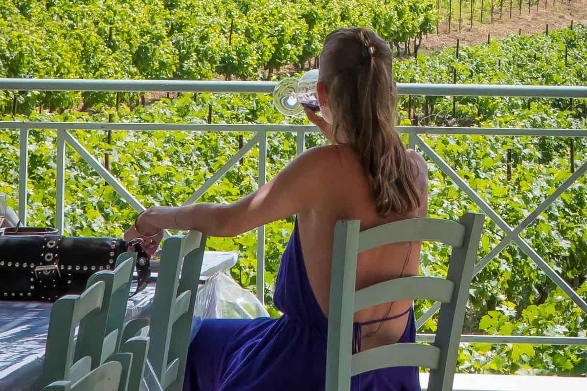 A Rhodes wine tour guest sips white wine on a terrace above green vineyard rows