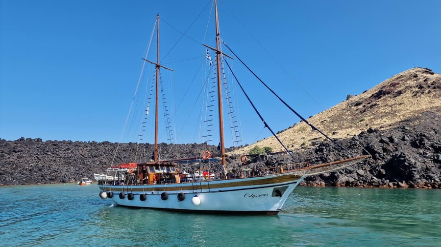 A two-masted wooden boat anchored in turquoise water below the dark lava of Nea Kameni on a Santorini boat trip