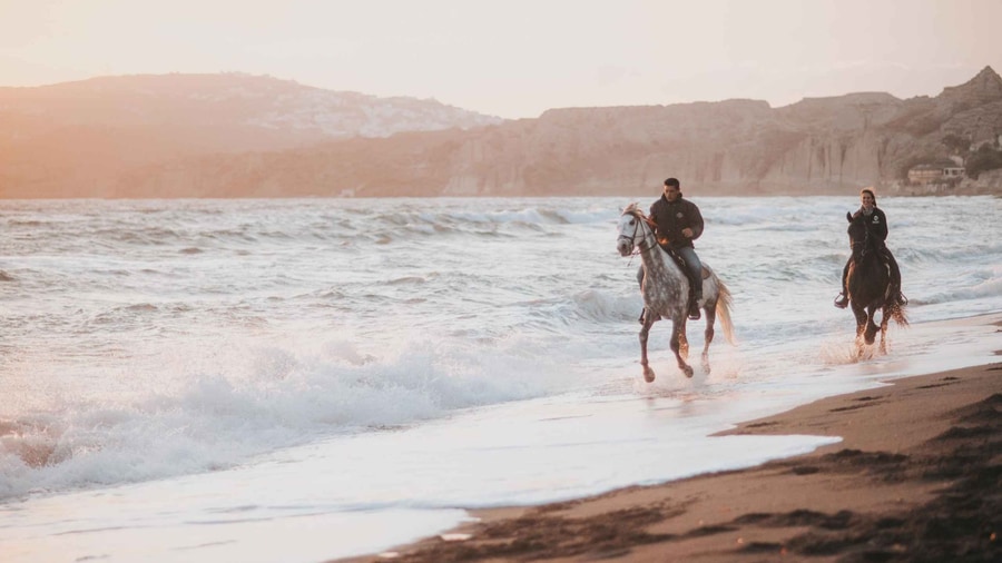 Santorini horse riding on a black sand beach, where two riders splash through the surf below the cliffs of the south coast
