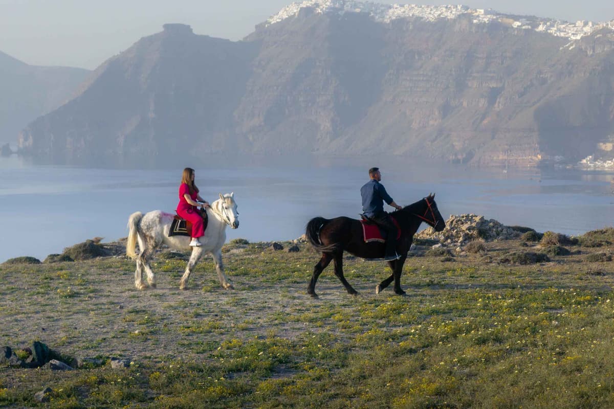 Santorini horse riding below the caldera, where two riders on a white and a black horse cross green grass above the sea with Fira on the cliffs