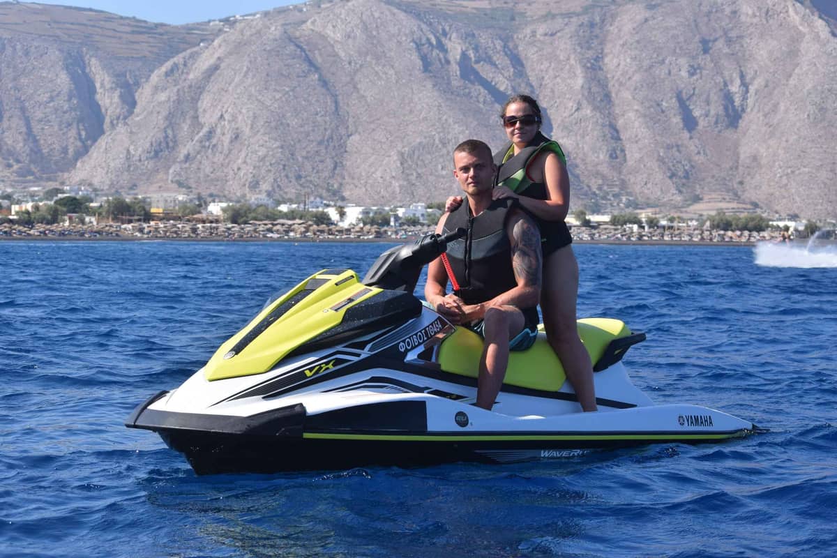 A Santorini jet ski with two riders on blue water below the bare mountain of the south coast