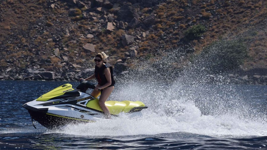 A Santorini jet ski rider in a life jacket throws up spray beside dark volcanic rock