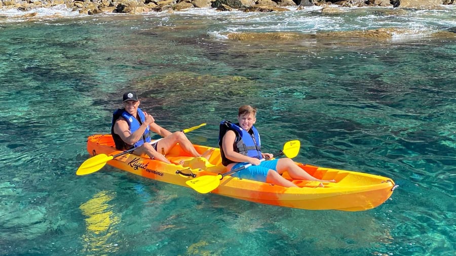 A Santorini kayak tour double sit-on-top kayak floats in clear green water beside a rocky volcanic shore