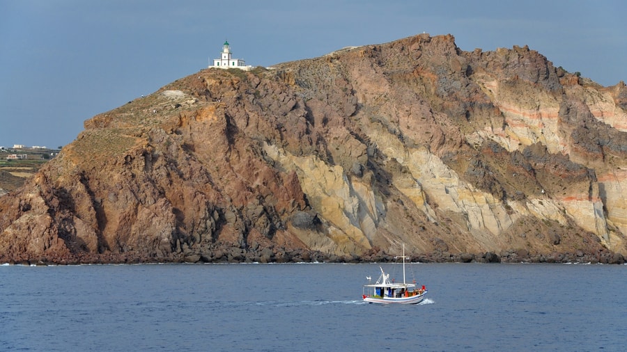 A Santorini sailing tour passes below the Akrotiri lighthouse on its cliff at the south-west tip of the island