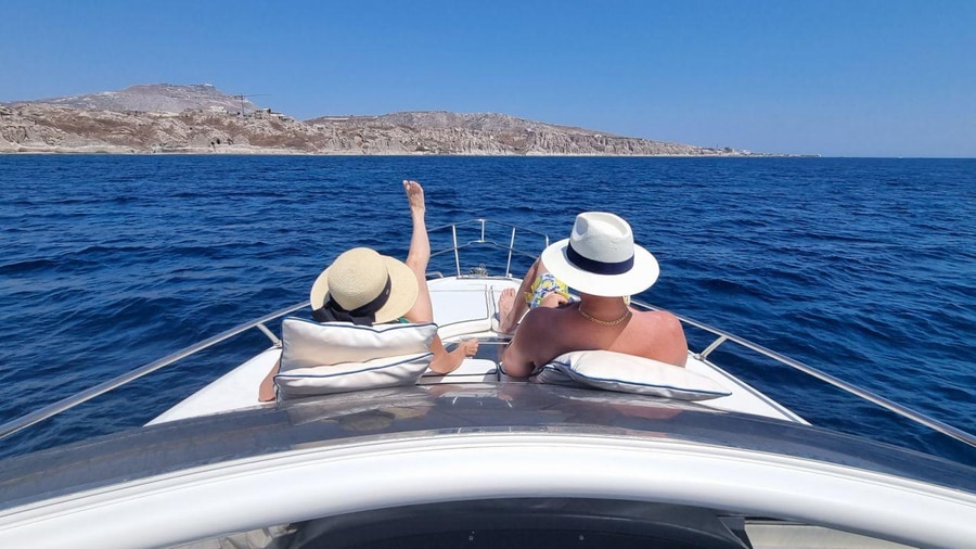 Two guests in sun hats relax on the bow of a Santorini yacht cruise over deep blue water