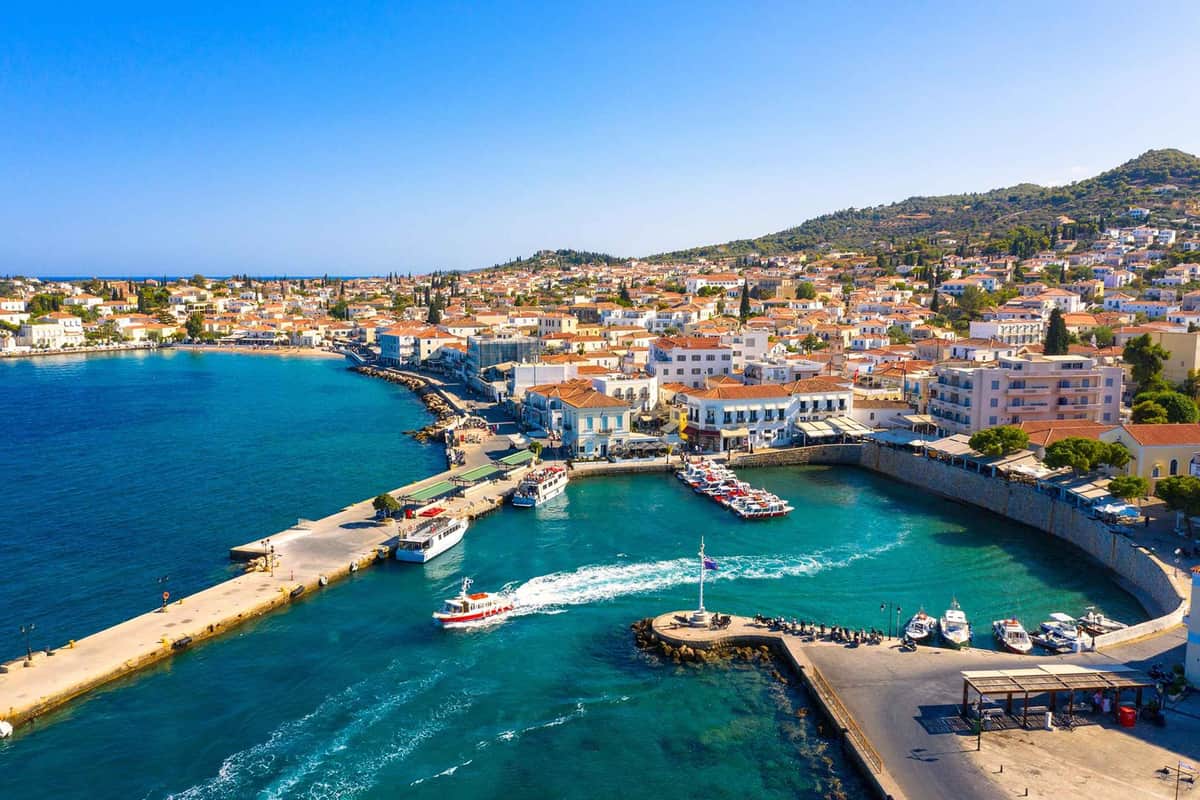 A Spetses day trip arrives at the main harbour of Spetses town, seen from above with the town along the shore and green hills behind