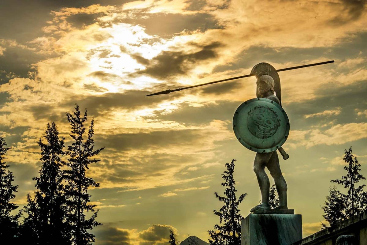A Thermopylae tour stands below the bronze statue of Leonidas with spear and shield against a golden evening sky