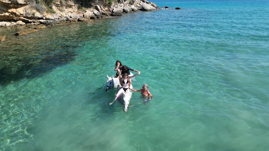 Riders sit on white horses that wade through the clear turquoise water of a rocky cove on Zakynthos horse riding
