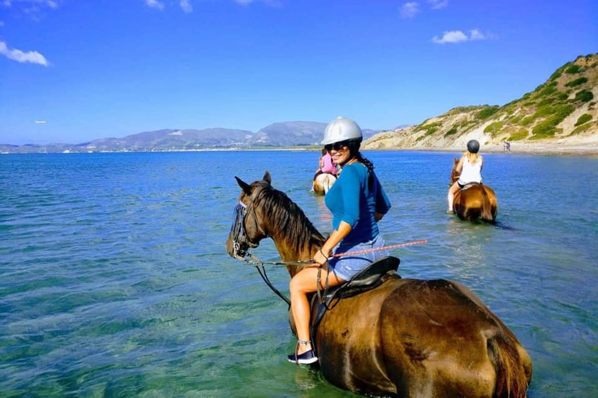 Zakynthos horse riding in the sea, with riders on brown horses wading through shallow blue water below green hills