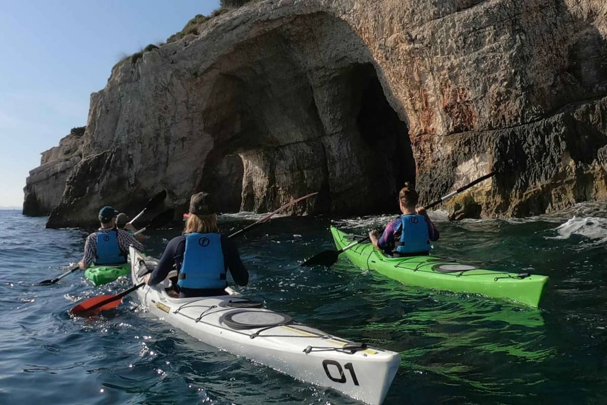 A Zakynthos kayak tour paddles three sea kayaks below a limestone cliff with a cave on the coast of Zakynthos