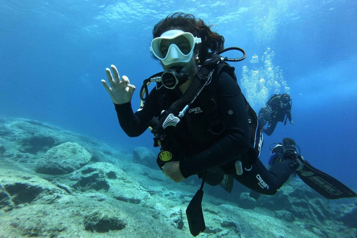 A Zakynthos scuba diving trip diver gives the OK sign above rocks in clear blue water